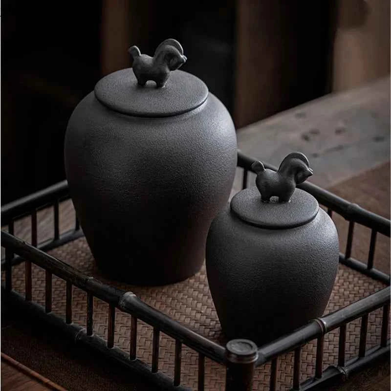 Black ceramic sealed tea storage jar with decorative animal-shaped lid handles on a woven tray