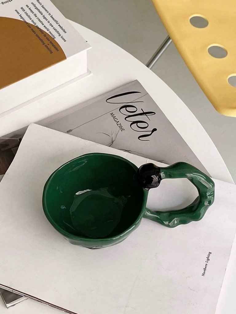 Green handmade ceramic coffee mug with unique textured handle on white magazine and book