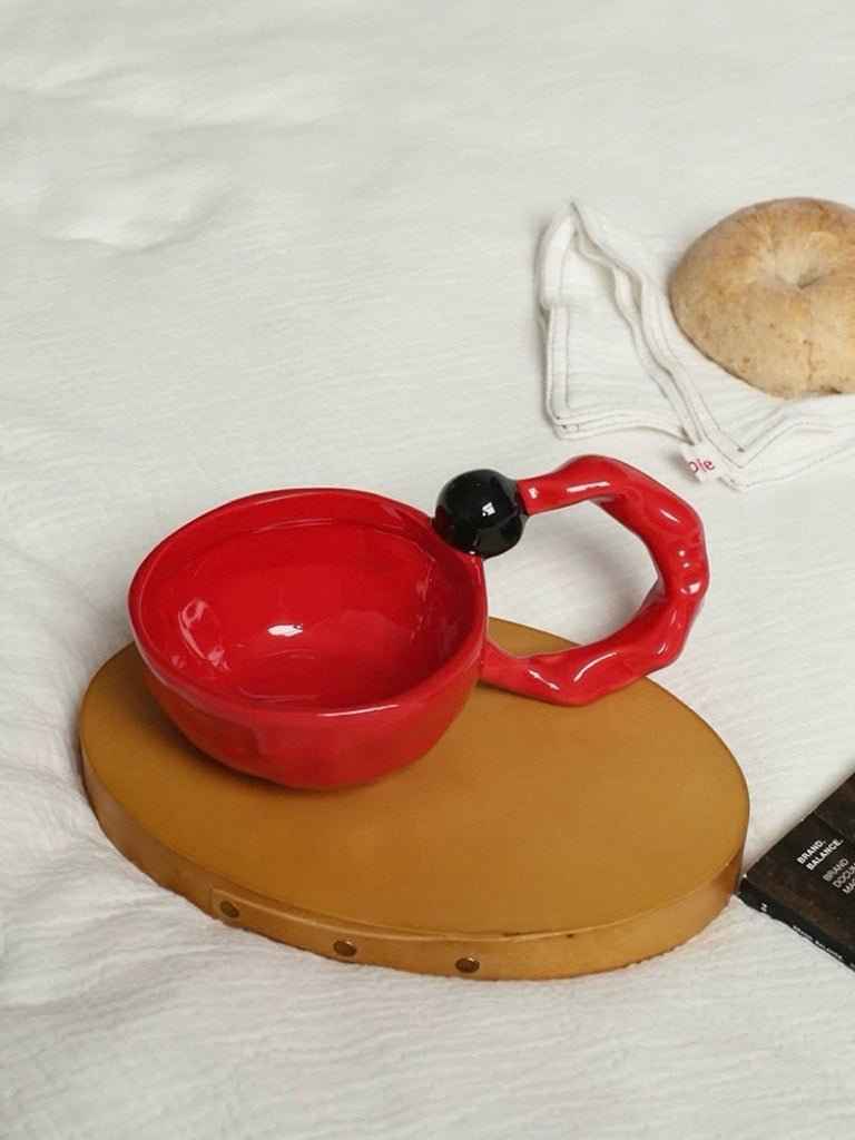 Red handmade ceramic coffee mugs with unique twisted handle design on a brown coaster