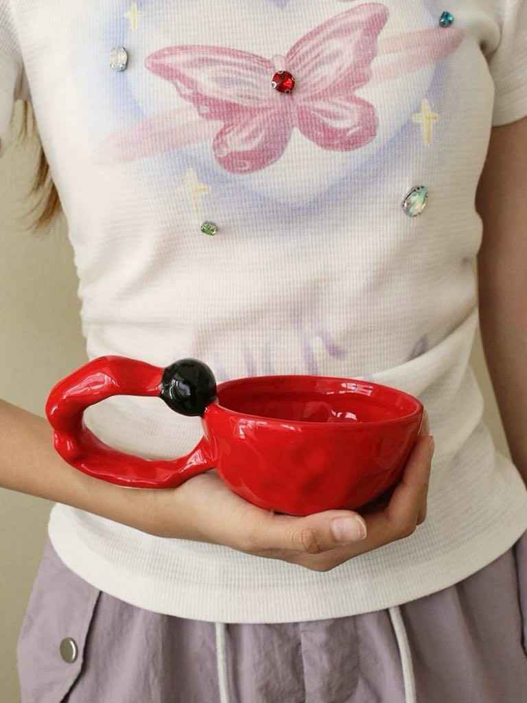 Hand holding a unique red handmade ceramic coffee mug with an artistic black bead handle